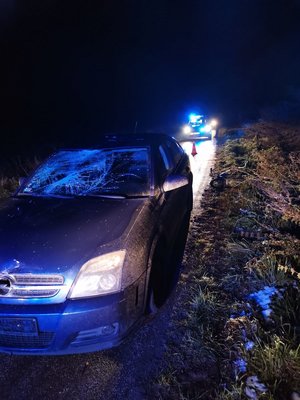 zmrok, niebieskie auto na jezdni z rozbitą przednią szybą, w tle radiowóz policji z włączonymi niebieskimi światłami, na poboczu leży rower.