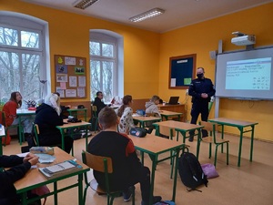 Policjant w klasie, dzieci w ławkach, funkcjonariusz stoi przed nimi, przedstawia prezentację multimedialną