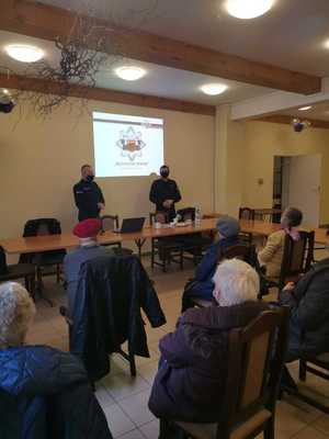 Na zdjęciach sala konferencyjna, na krzesłach grupa seniorów przysłuchuje się prezentacji przedstawianej przez umundurowanego policjanta oraz strażaka. Na ścianie wyświetlana jest prezentacja multimedialna.